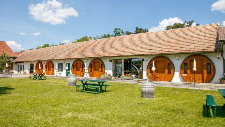 Ein langes Geb&auml;ude mit runden Holzt&uuml;ren, umgeben von einer gr&uuml;nen Wiese und Picknicktischen.