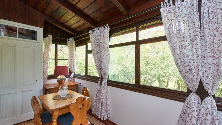 Ein gem&uuml;tlicher Wintergarten mit Holzm&ouml;beln, floralen Vorh&auml;ngen und Blick ins Gr&uuml;ne.