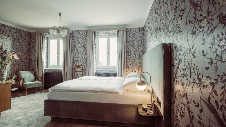 Elegantes Schlafzimmer mit floral gemusterten Tapeten, gro&szlig;em Bett und zwei Fenstern.