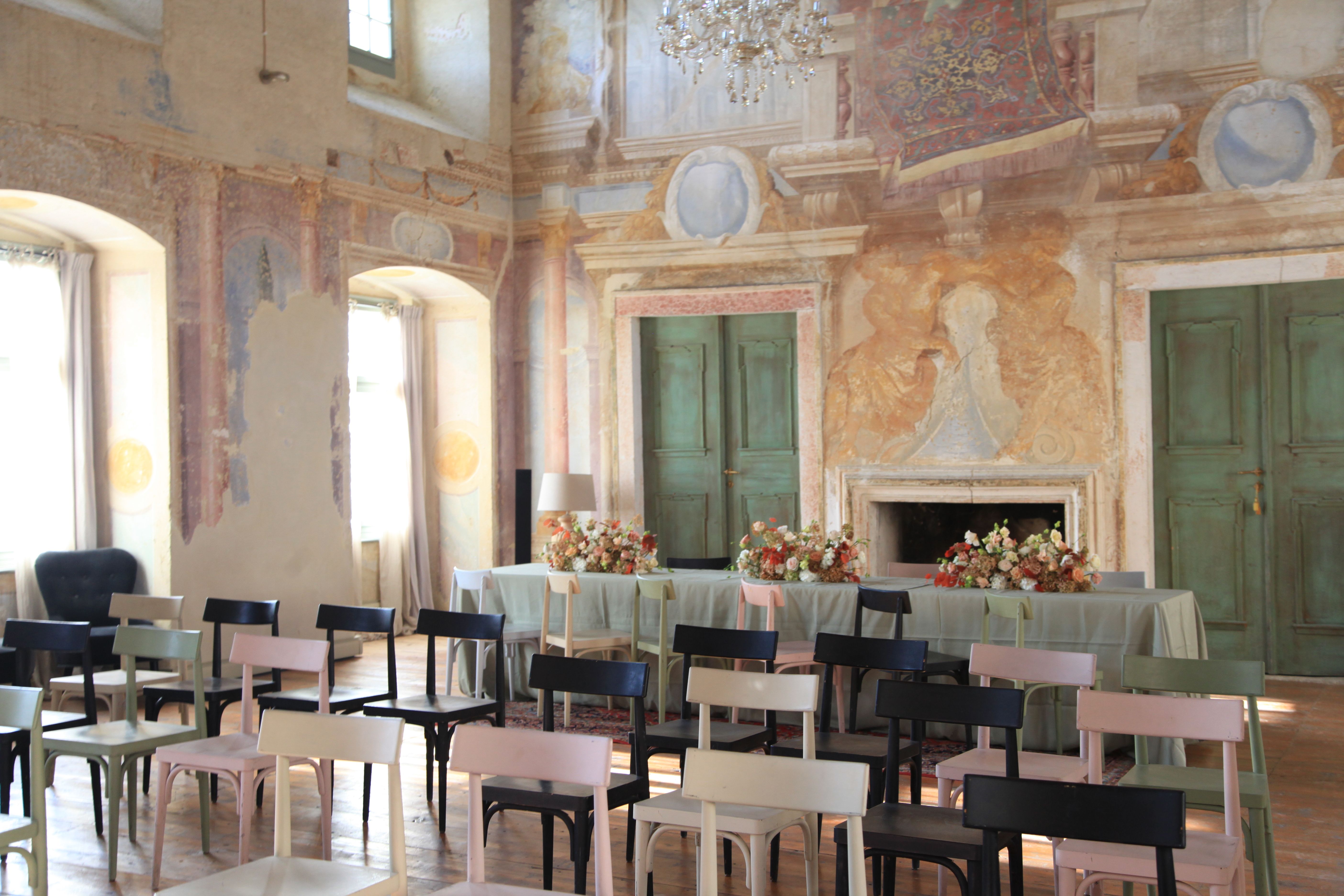 Ein festlich dekorierter Saal mit bemalten Wänden, Stühlen und einem Tisch mit Blumen.
