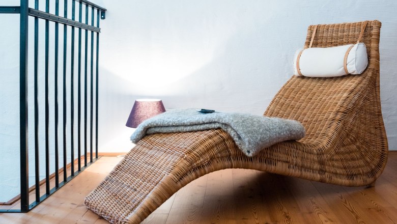 Eine gem&uuml;tliche Leseecke mit einem geflochtenen Liegestuhl, einer Decke und einer Lampe auf Holzboden.