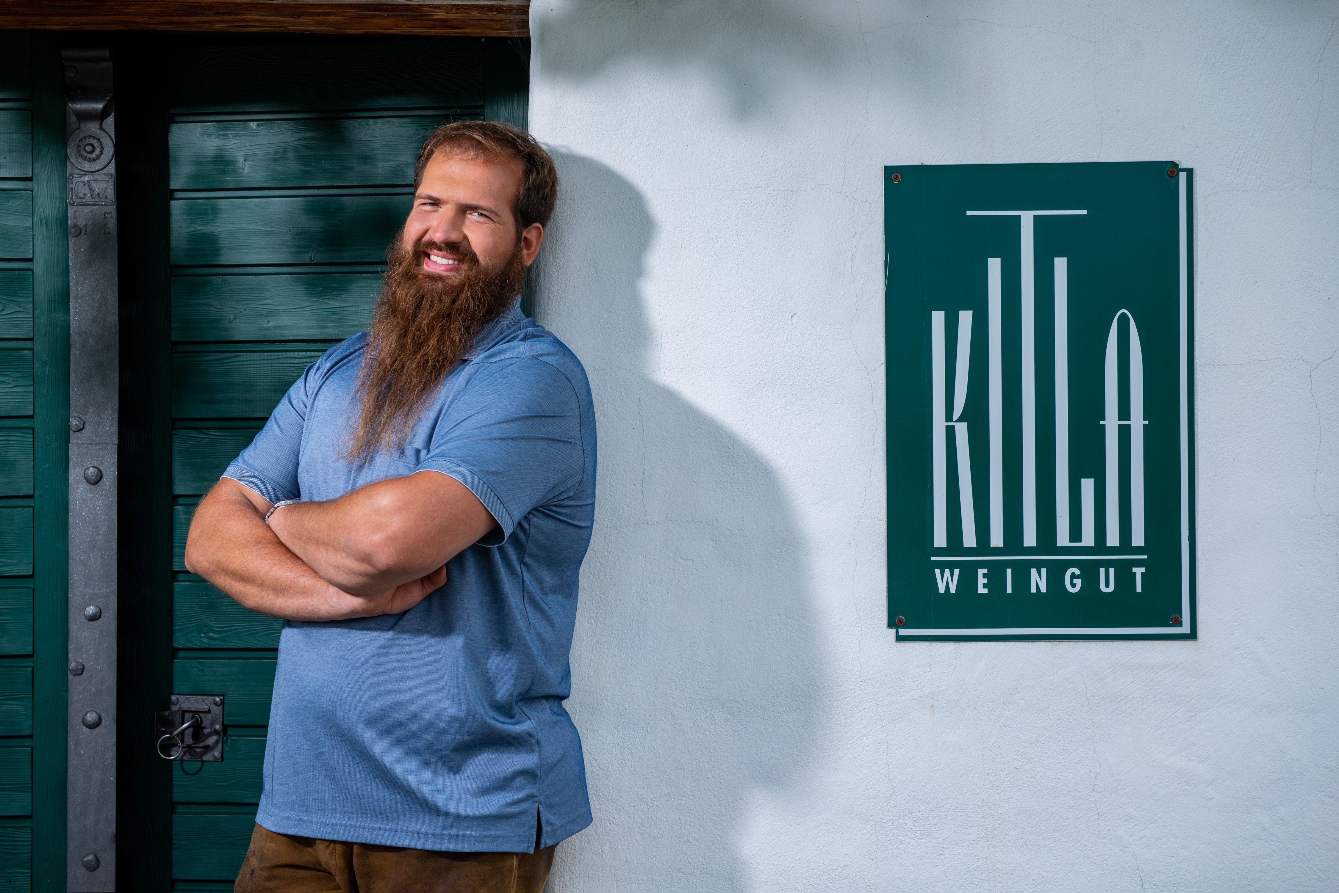 Ein Mann mit Bart steht lächelnd vor einem Schild mit der Aufschrift 'Kitla Weingut'.