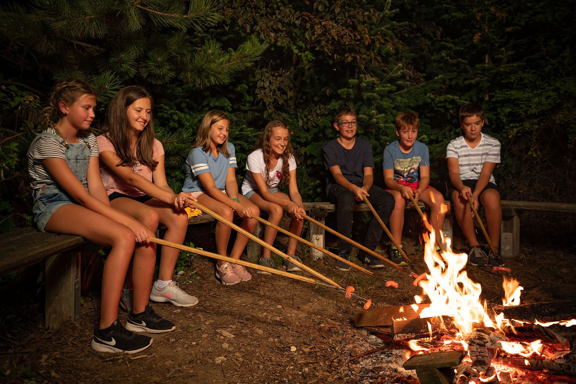 Kinder sitzen um ein Lagerfeuer und grillen Würstchen an langen Stöcken.