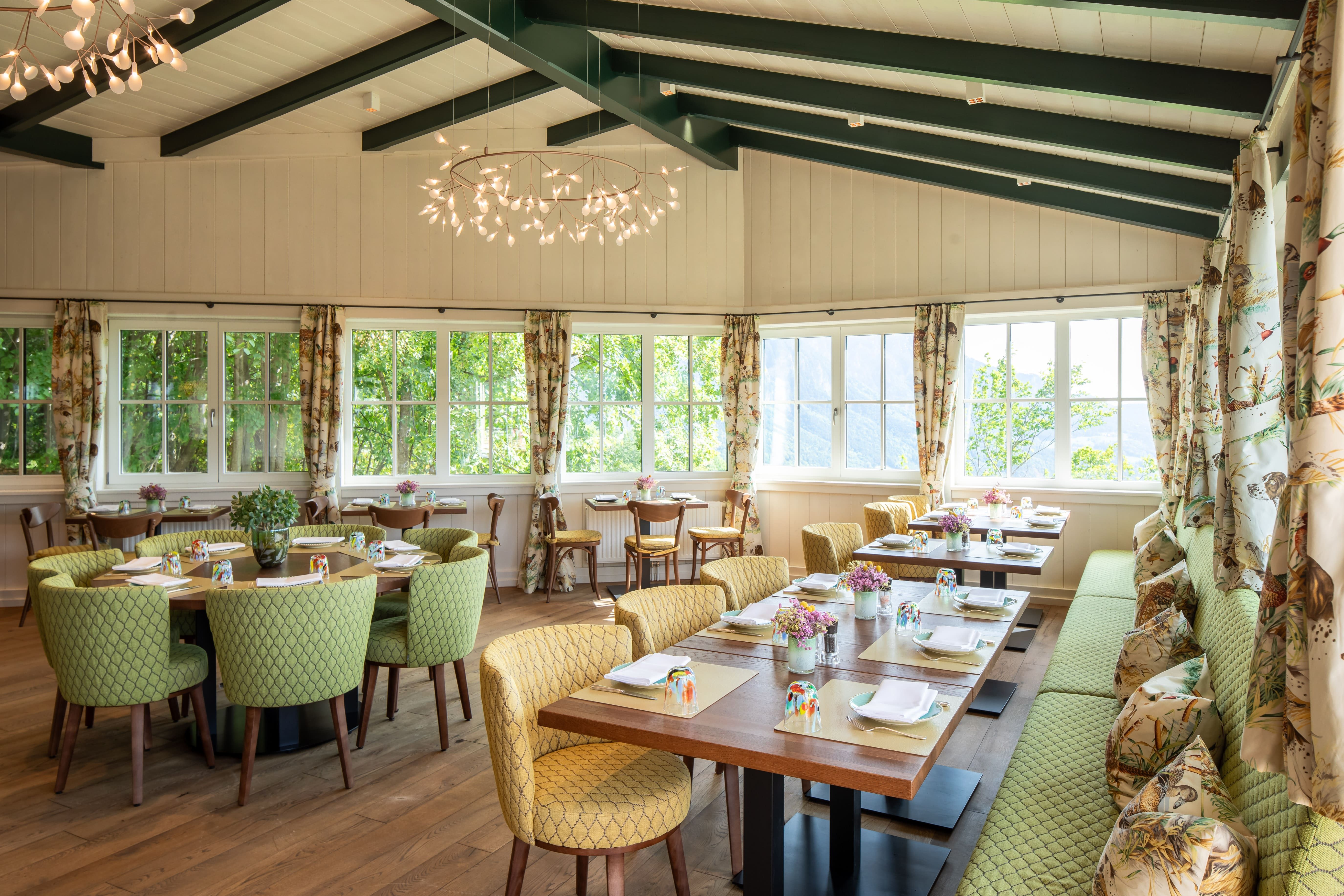 Ein heller Wintergarten mit gedeckten Tischen, grünen Stühlen und großen Fenstern.