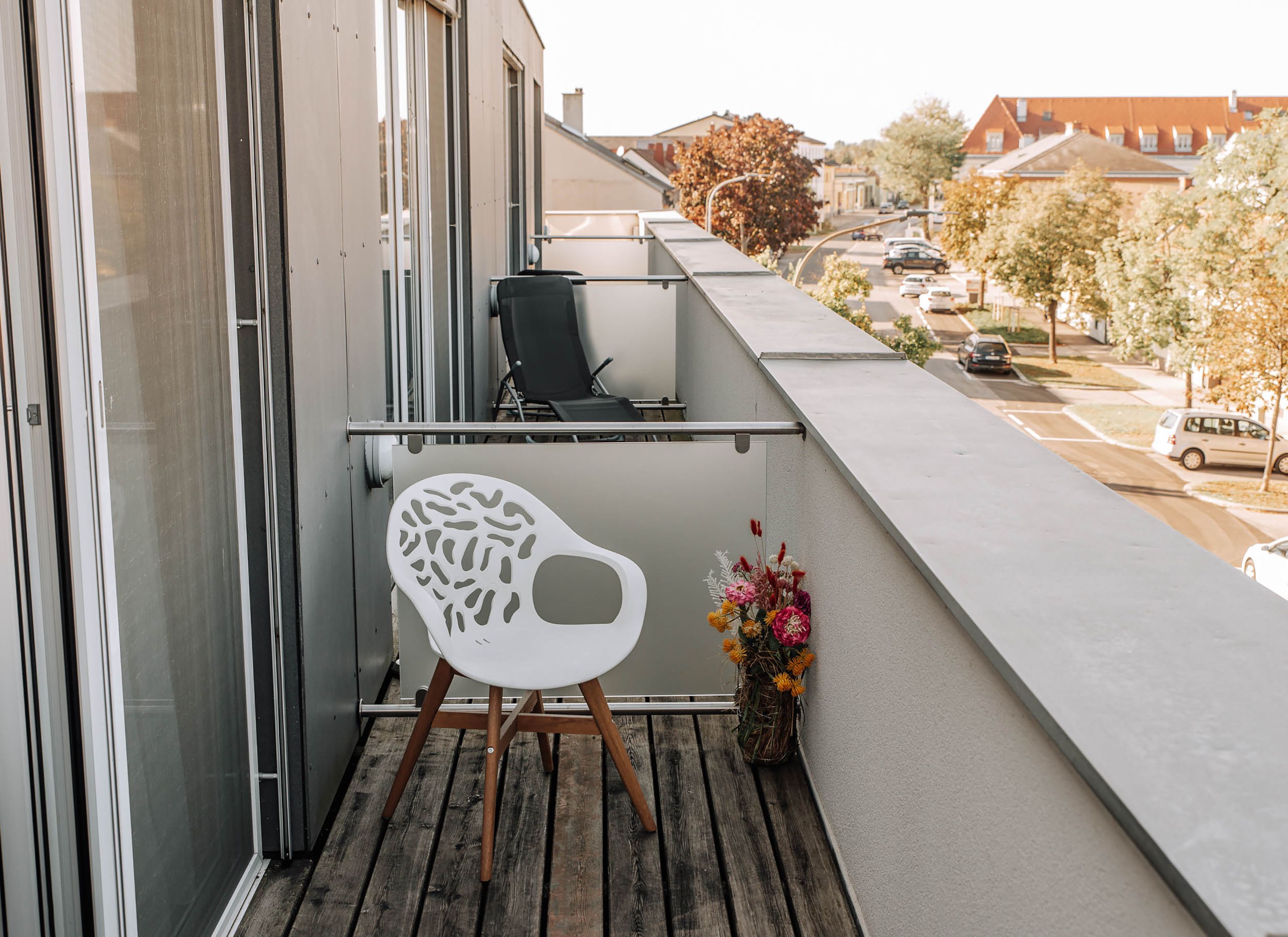 Balkon mit weißem Stuhl und Blumenstrauß, Blick auf Straße und Gebäude.