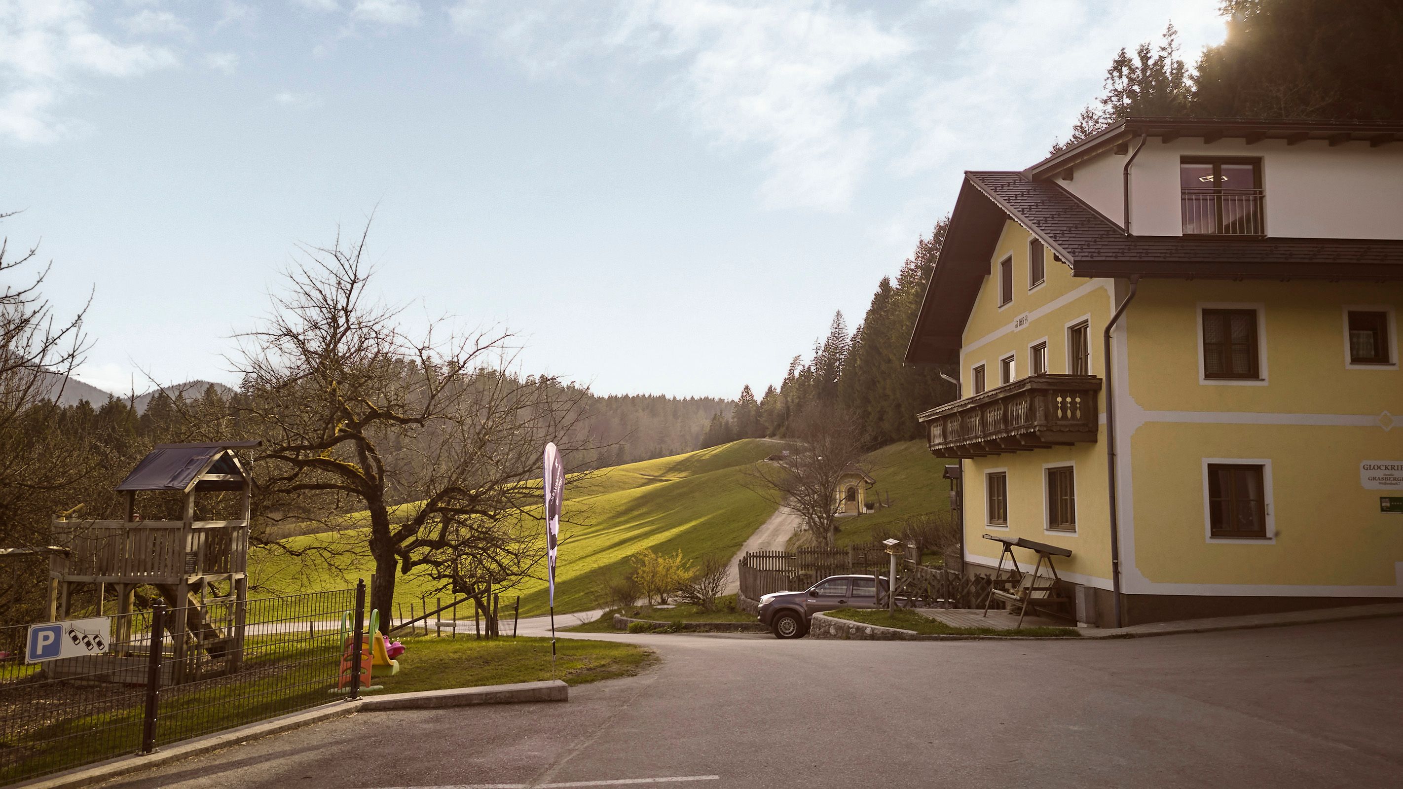 Ein gelbes Haus mit Balkon steht neben einem Spielplatz und einer Straße, die durch grüne Hügel führt. Der Himmel ist klar und die Sonne scheint.
