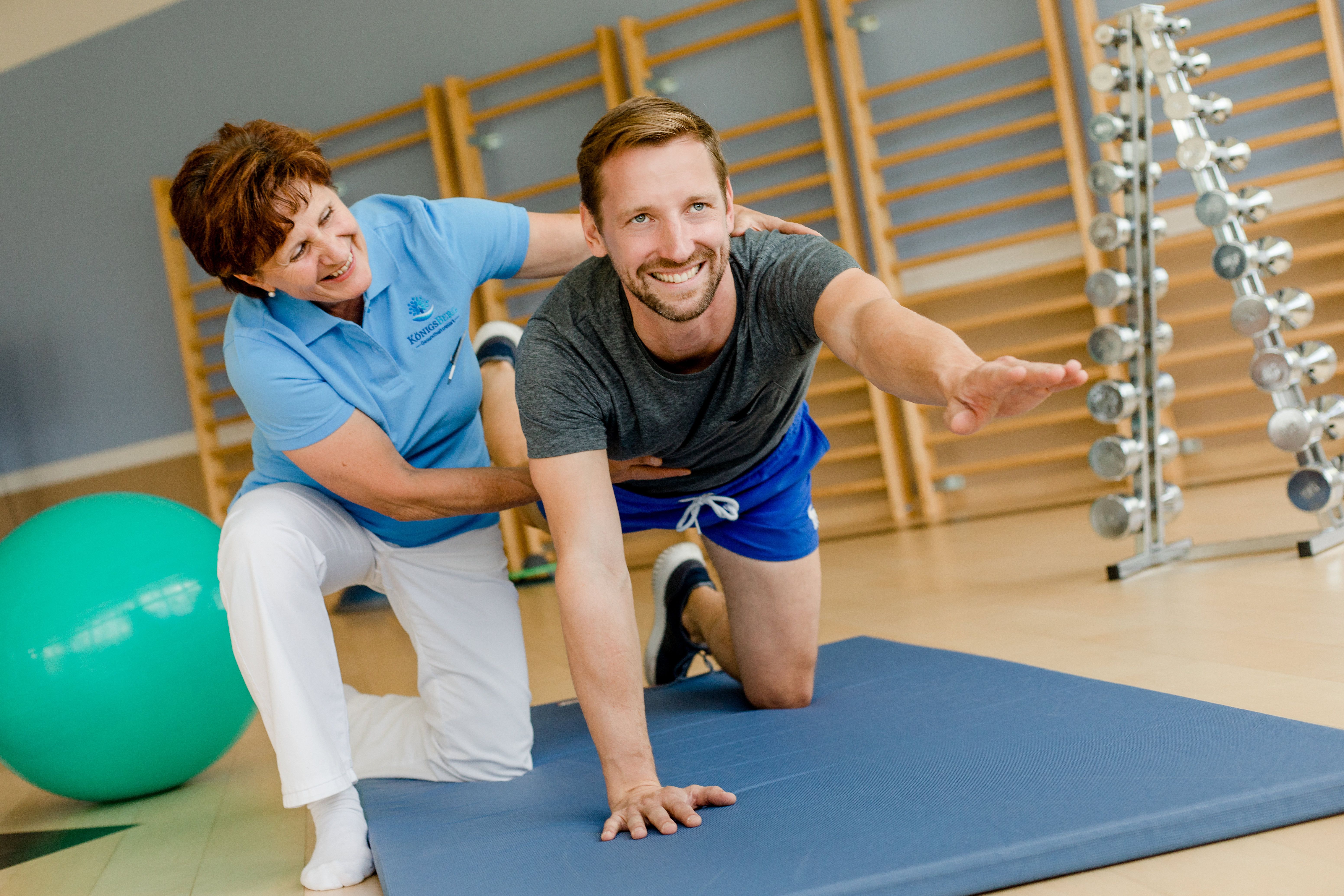 Ein Mann macht eine Übung auf einer Matte, unterstützt von einer Therapeutin in einem Fitnessraum.