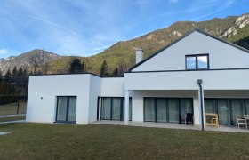 Modernes weißes Haus mit großen Fenstern vor einer Bergkulisse.