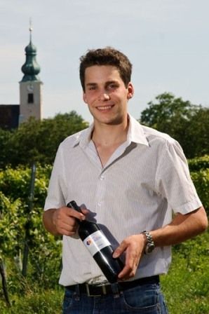 Ein Mann steht in einem Weinberg und hält eine Weinflasche. Im Hintergrund ist ein Kirchturm zu sehen.
