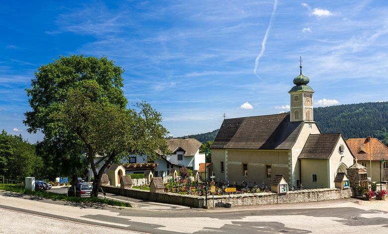 Kleine Kirche mit Turm in einem Dorf, umgeben von Bäumen und Häusern, unter blauem Himmel.