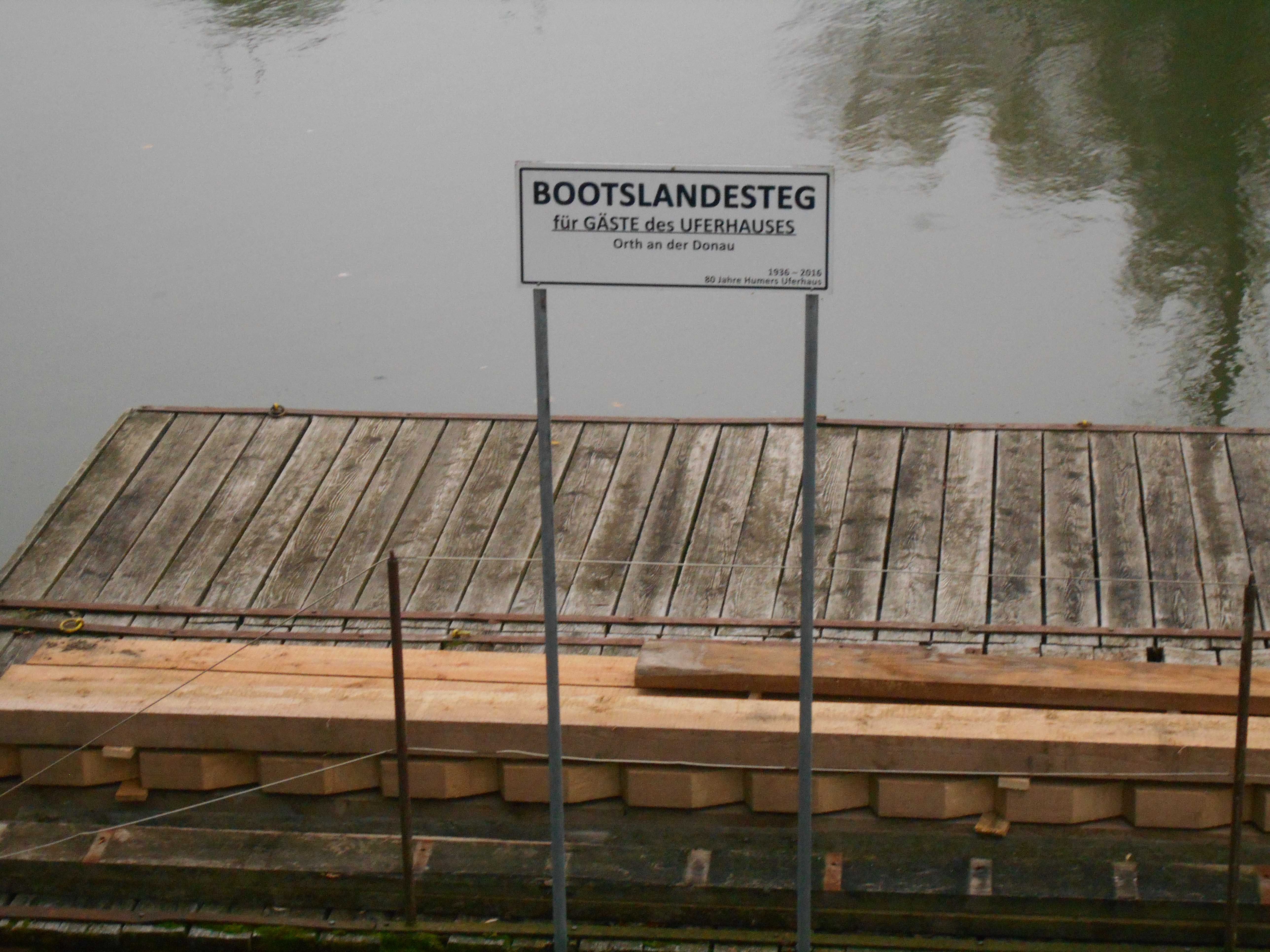 Holzsteg mit Schild 'Bootslandesteg für Gäste des Uferhauses' an einem ruhigen Gewässer.
