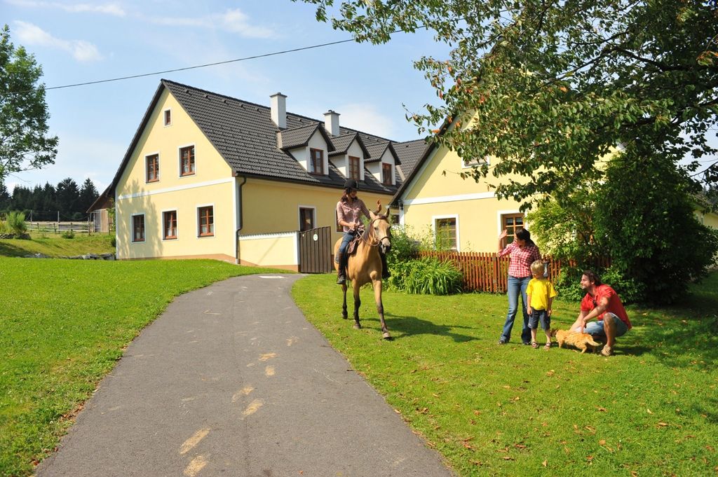 Ein Reiter auf einem Pferd nähert sich einer Familie vor einem gelben Haus auf einem Bauernhof.