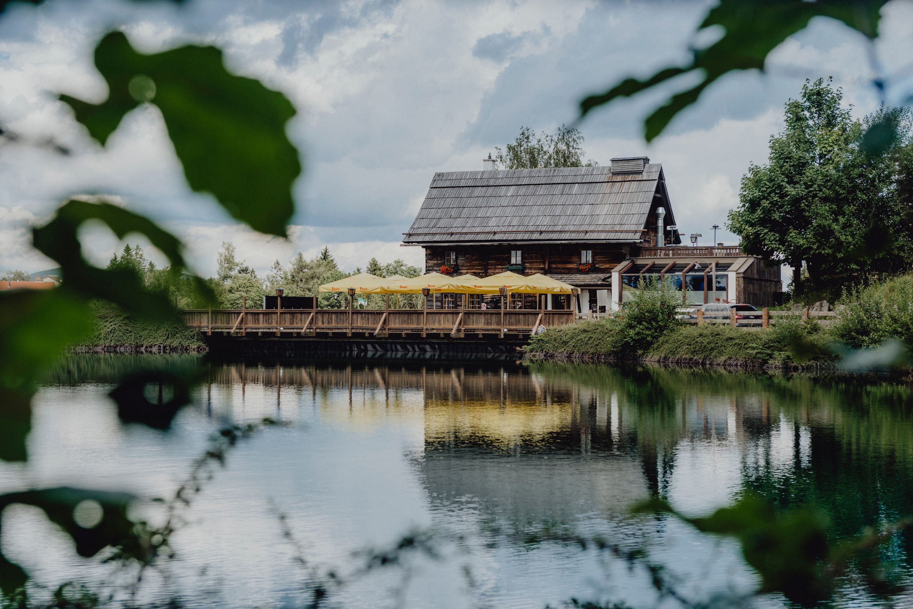 Ein rustikales Holzhaus mit gelben Sonnenschirmen spiegelt sich in einem ruhigen See, umgeben von grüner Natur.