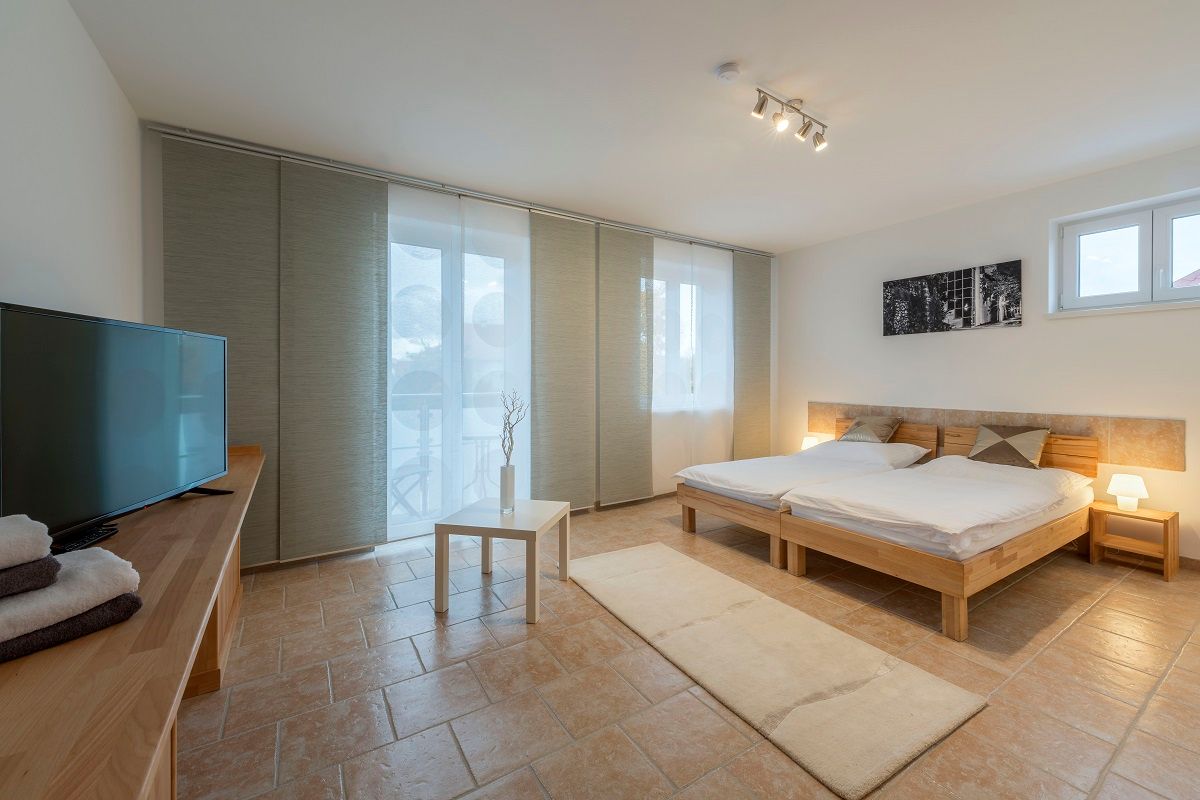 Modernes Schlafzimmer mit Doppelbett, Fernseher, Tisch und großen Fenstern mit Vorhängen.