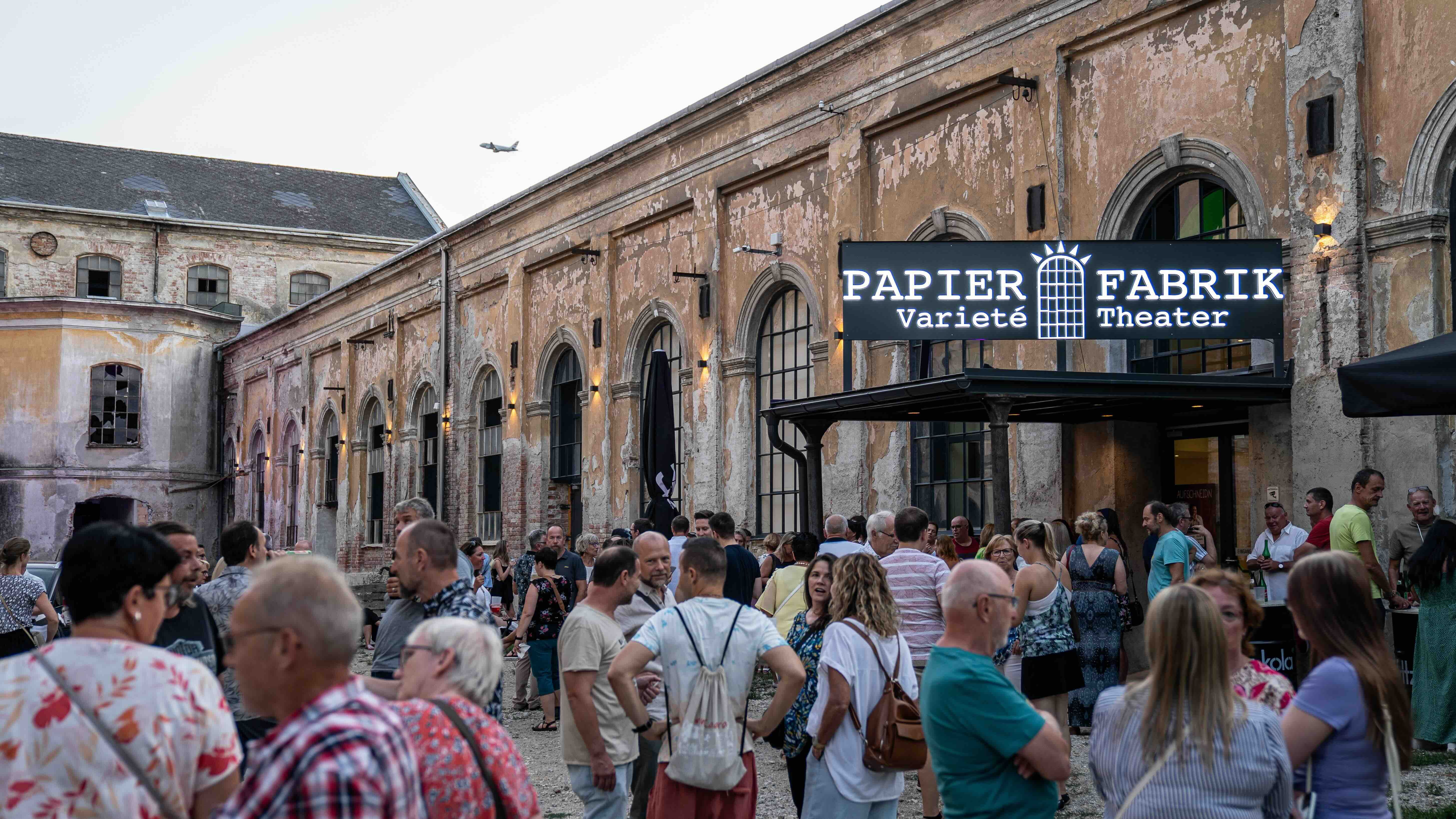 Menschenmenge vor einer alten Papierfabrik mit einem Schild 'Papier Fabrik Varieté Theater'.