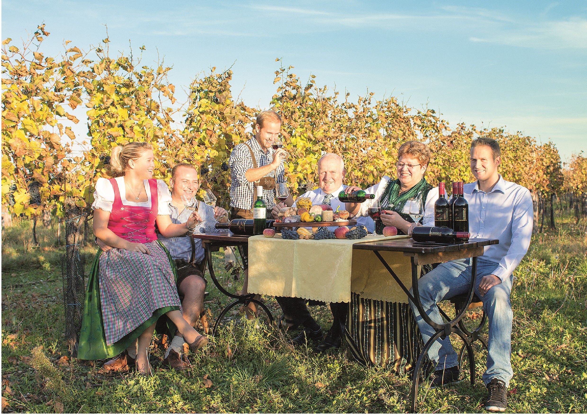 Eine Familie sitzt an einem Tisch im Weinberg und genießt Wein und Snacks.