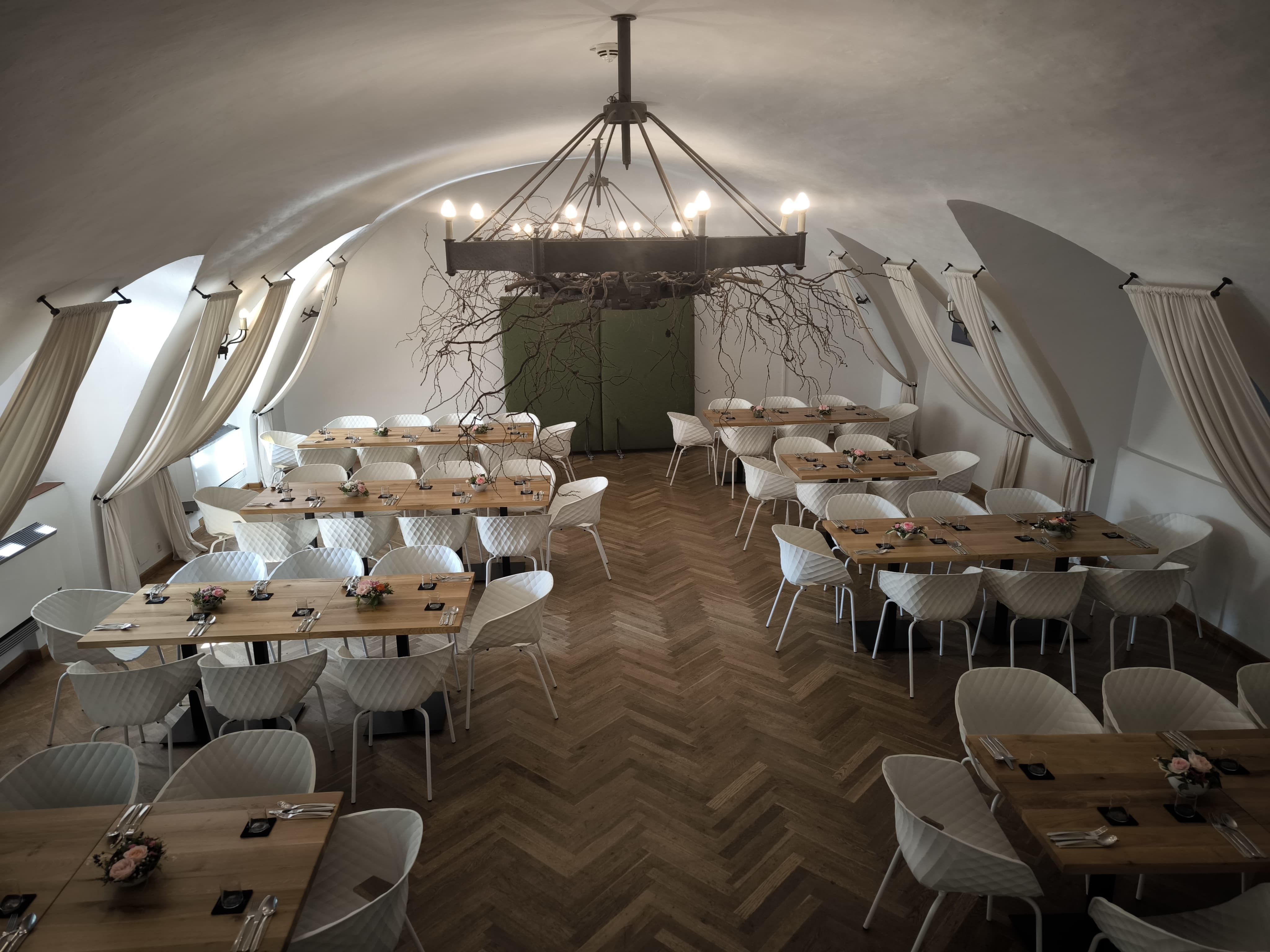 Eleganter Saal mit Holztischen, Parkettboden, weißen Stühlen und eisnerner Kronleuchter.