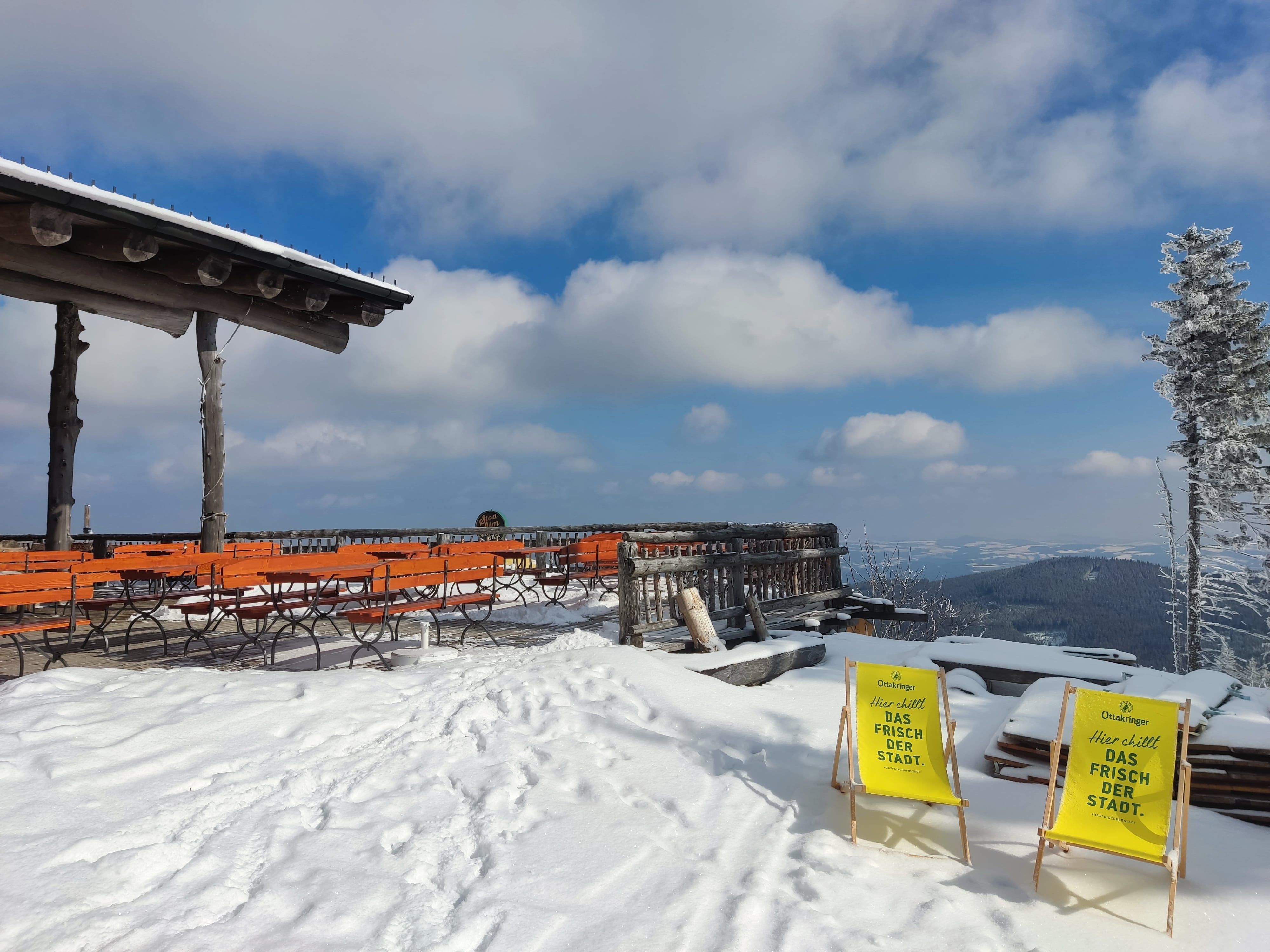 Verschneite Terrasse mit orangefarbenen Bänken und zwei gelben Liegestühlen, umgeben von schneebedeckten Bäumen und Bergen im Hintergrund.