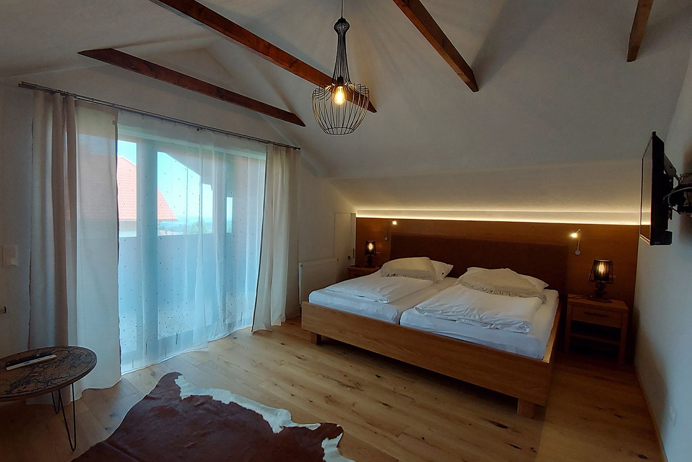 Ein gemütliches Doppelzimmer mit Holzboden, einem großen Bett, Vorhängen und einem kleinen Tisch. Eine Lampe hängt von der Decke.