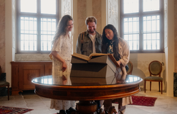 Drei Personen stehen in einem historischen Raum um einen runden Tisch und betrachten ein aufgeschlagenes Buch; der Raum ist mit hohen Fenstern, Stuckdecken und einem roten Teppich ausgestattet.