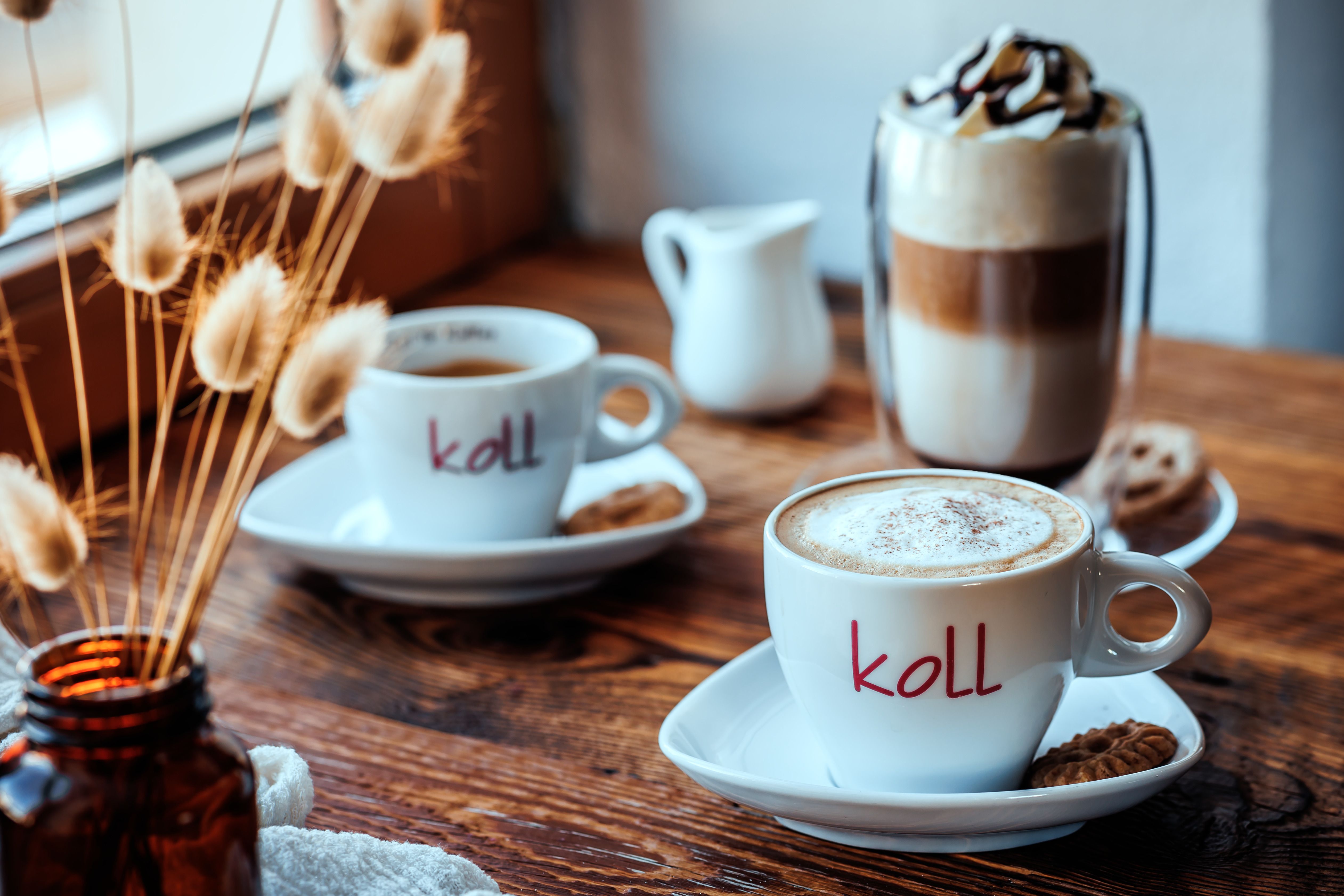 Zwei Tassen Kaffee auf einem Holztisch, eine mit Milchschaum, daneben ein Glas Latte Macchiato und ein Krug.