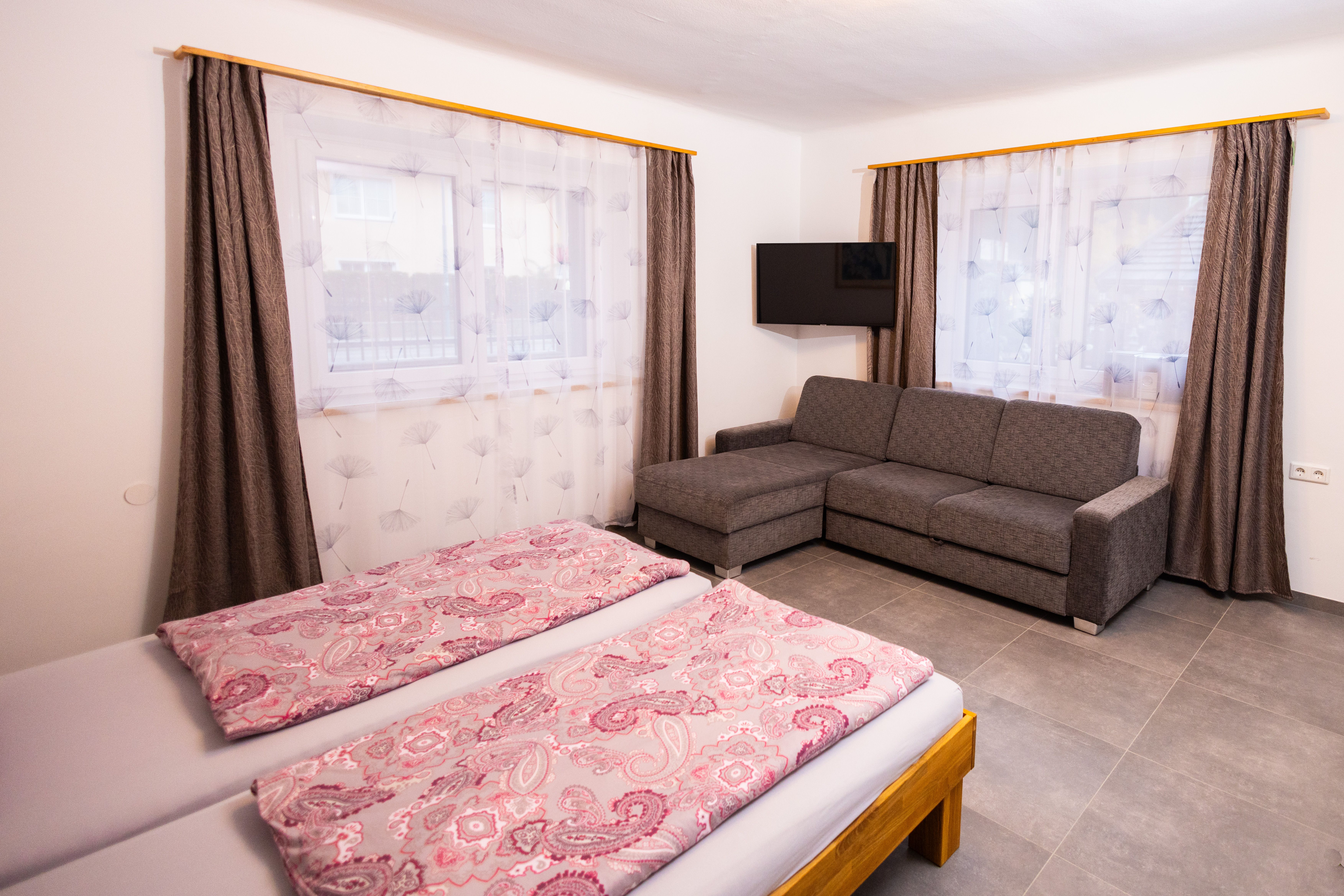 Ein Schlafzimmer mit Doppelbett, grauem Sofa, Fernseher und zwei Fenstern mit Vorhängen.