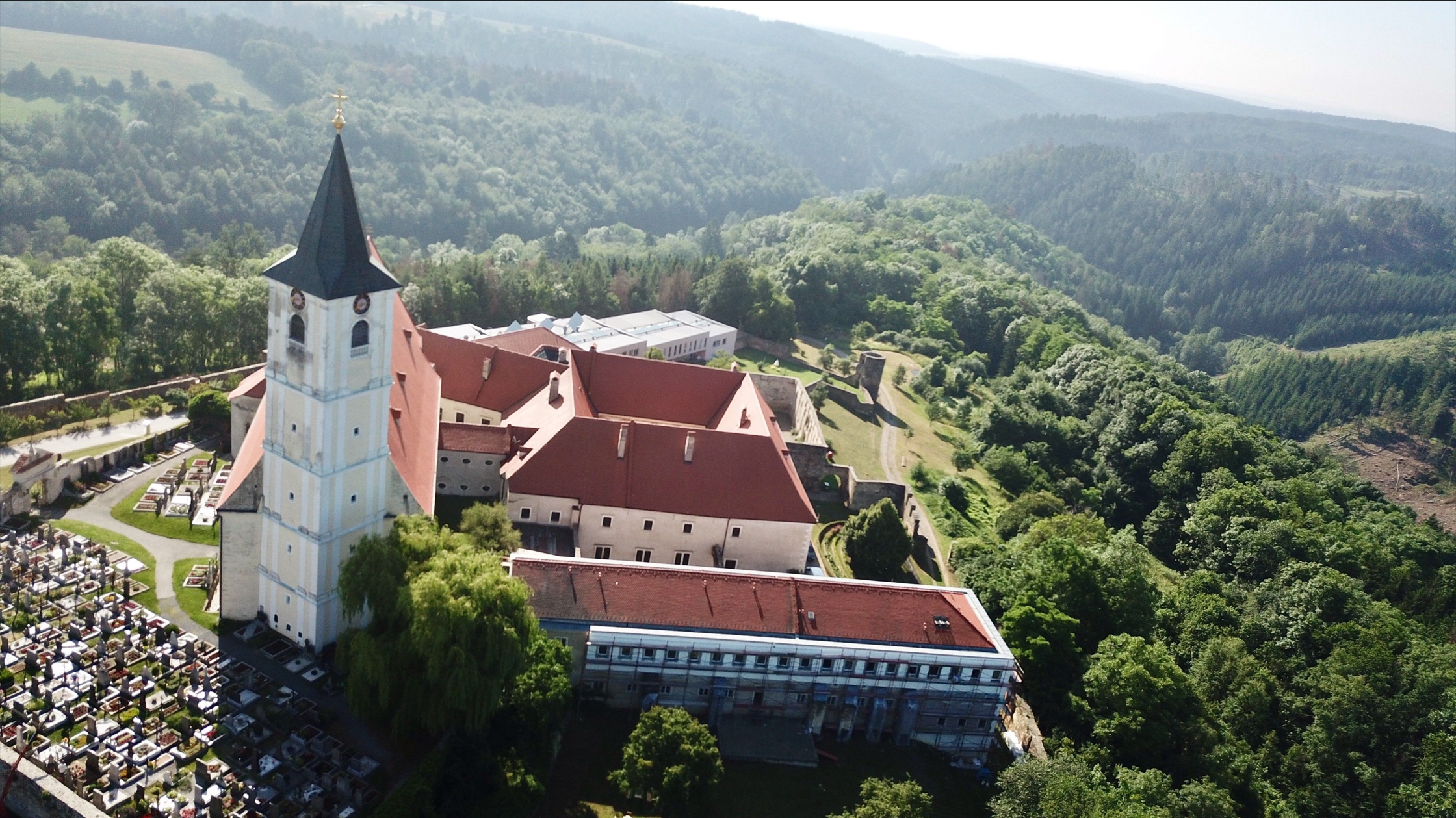 Luftaufnahme eines Klosters mit rotem Dach und einem Friedhof daneben, umgeben von bewaldeten Hügeln.