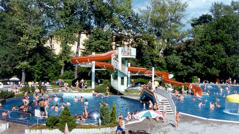 Ein Freibad mit vielen Menschen, einer Wasserrutsche und Bäumen im Hintergrund.