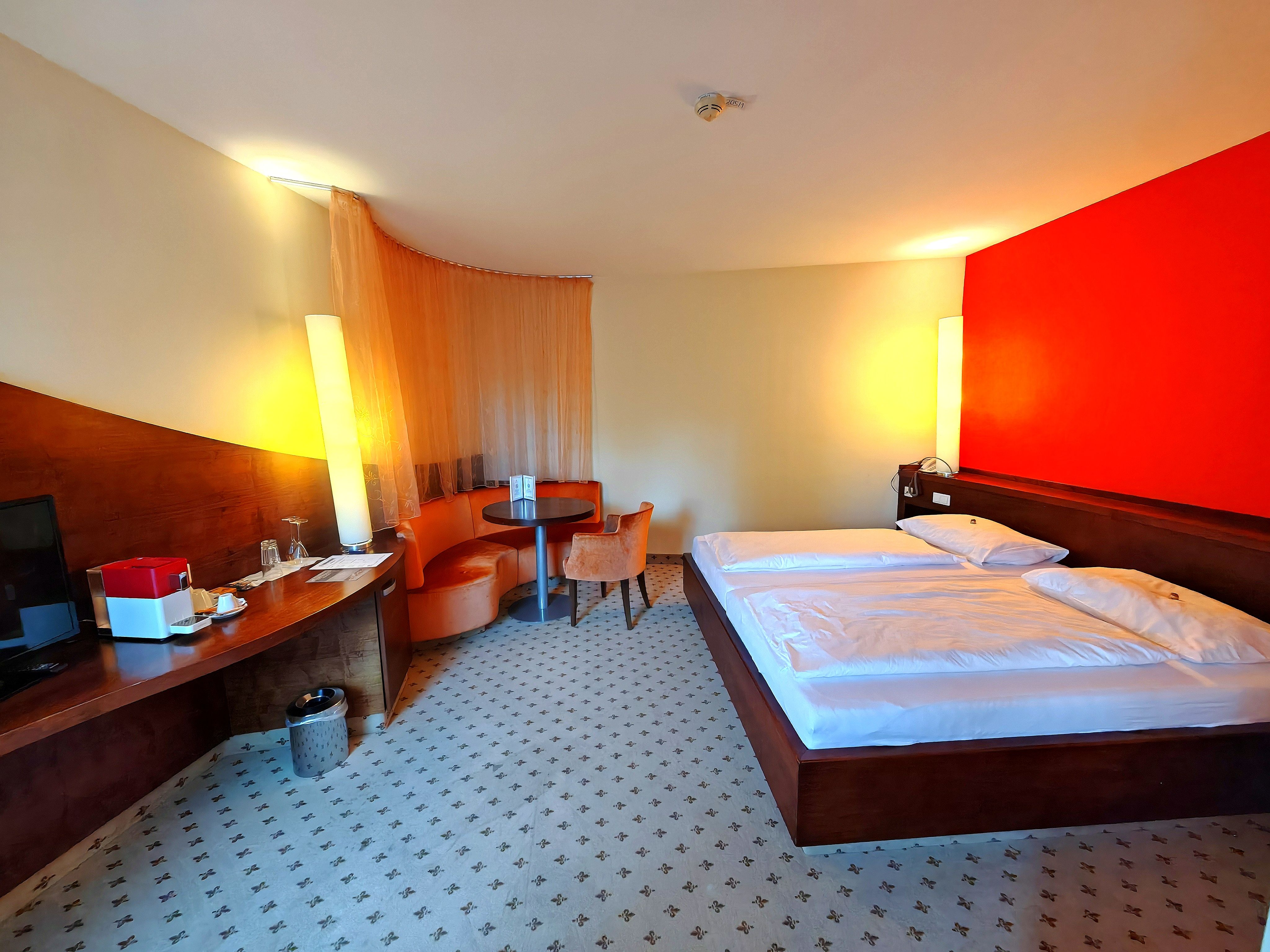 Hotelzimmer mit Doppelbett, roter Akzentwand, Schreibtisch und Sitzbereich.