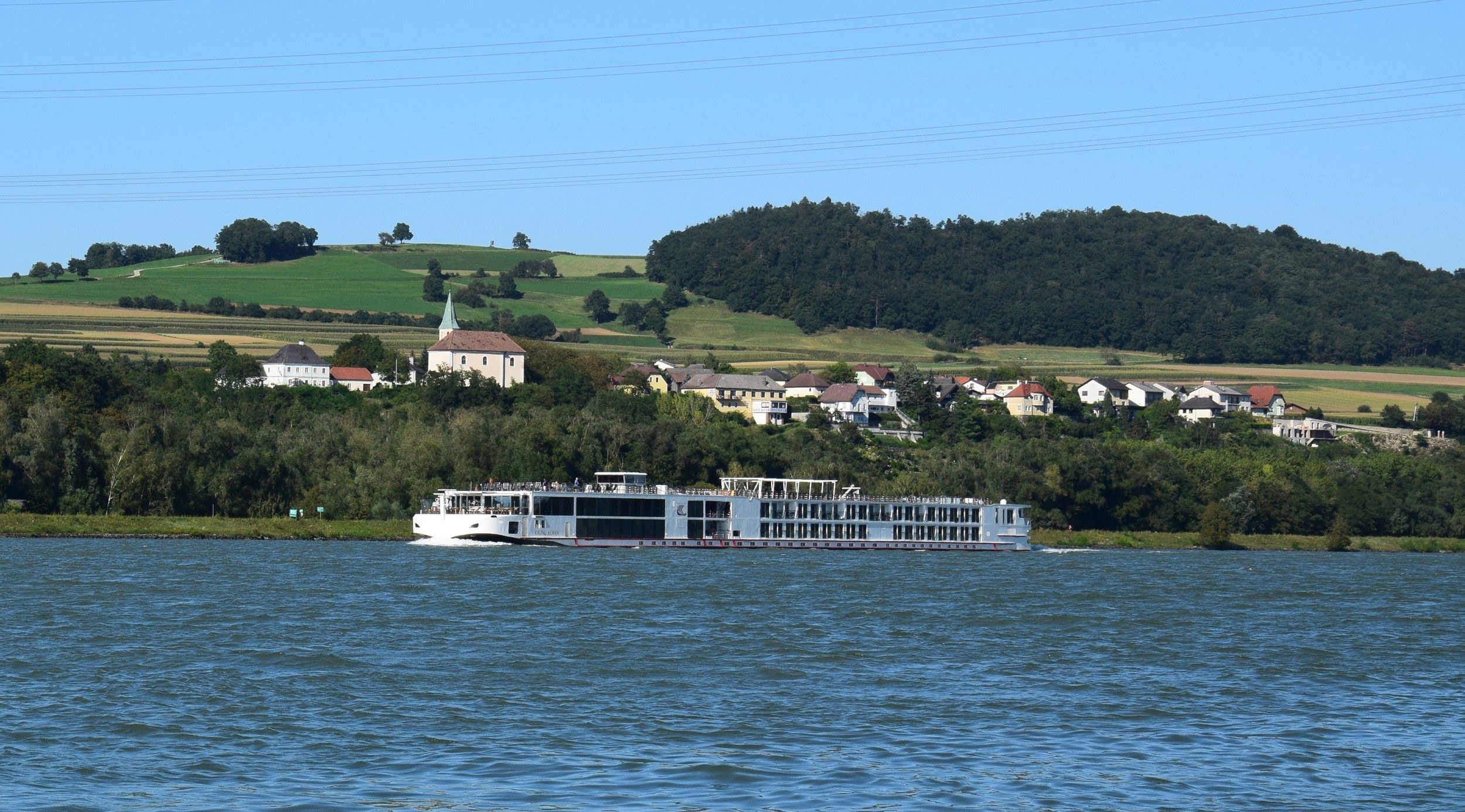 Ein Flusskreuzfahrtschiff fährt auf einem Fluss vor einer grünen Hügellandschaft mit einem Dorf im Hintergrund.