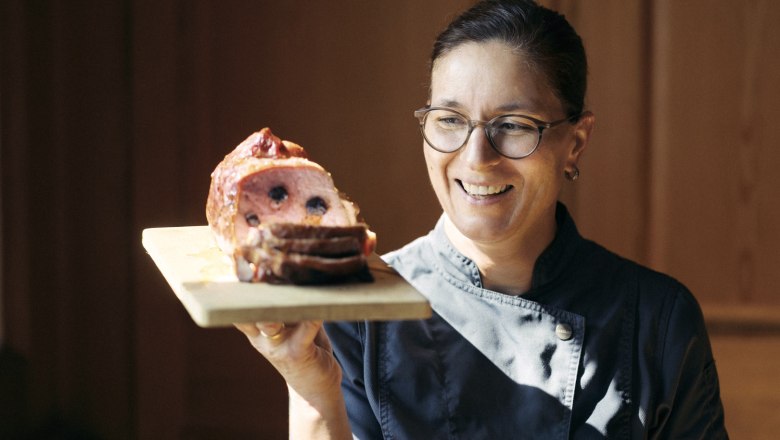 Eine Frau in Kochkleidung hält ein Holzbrett mit einem aufgeschnittenen Fleischgericht.