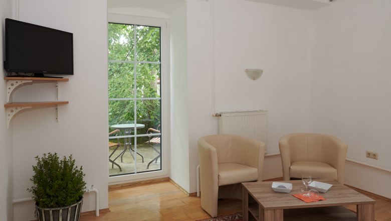 Wohnzimmer mit zwei beigen Sesseln, Holztisch, Fernseher und Balkon mit Tisch und St&uuml;hlen.