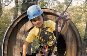 Ein Kind mit Helm und Kletterausr&uuml;stung in einem h&ouml;lzernen Kletterpark-Tunnel.