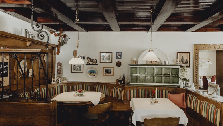 Gem&uuml;tliche Gaststube mit Holzm&ouml;beln, Kachelofen und dekorativen Bildern an der Wand.