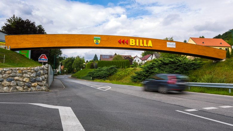 Eine Holzbr&uuml;cke mit dem Schriftzug 'BILLA' &uuml;berspannt eine Stra&szlig;e in einer l&auml;ndlichen Umgebung.