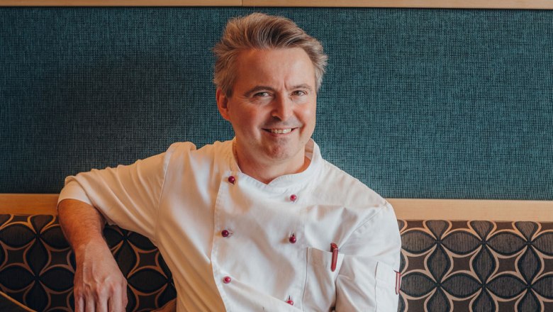 Ein lächelnder Mann in weißer Kochjacke sitzt auf einer gepolsterten Bank vor einer gemusterten Wand.