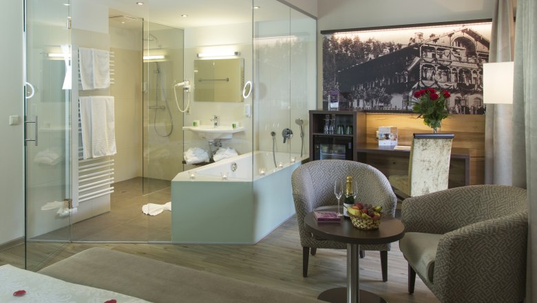 Luxuri&ouml;ses Hotelzimmer mit Glasbad, gem&uuml;tlichen Sesseln, Tisch mit Champagner und Fr&uuml;chten, sowie einem gro&szlig;en Bett mit Rosenbl&auml;ttern.