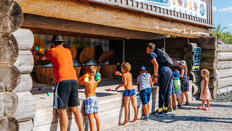 Kinder und Erwachsene werfen bunte B&auml;lle in einem Spielstand aus Holz im Freien.