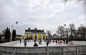 Menschen beim Eislaufen auf einem Freiluftplatz vor einem gelben Gebäude.