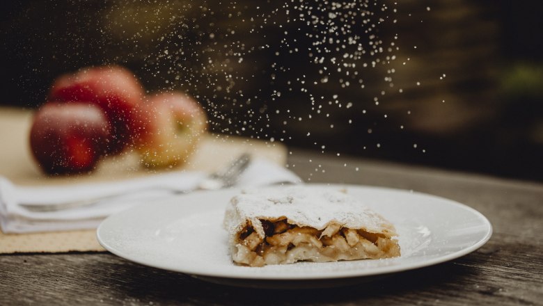 Apfelstrudel mit Puderzucker auf Teller, &Auml;pfel im Hintergrund.