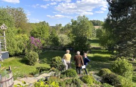 Drei Personen stehen in einem gr&uuml;nen Garten mit B&auml;umen und Str&auml;uchern, umgeben von einer l&auml;ndlichen Landschaft unter blauem Himmel mit Wolken.