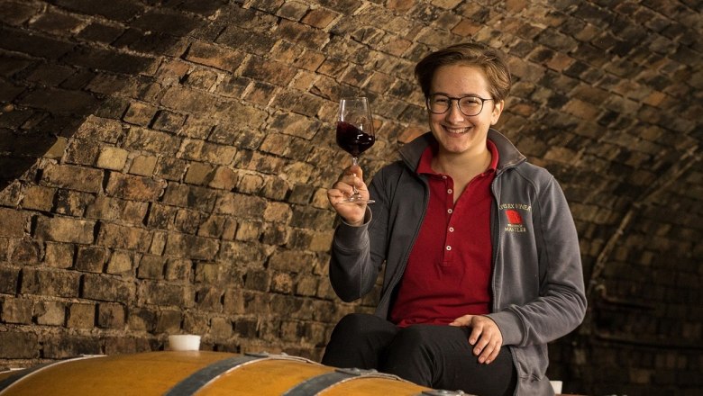 Eine Person sitzt auf einem Weinfass in einem Weinkeller und h&auml;lt ein Glas Rotwein in der Hand.
