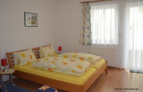 Ein gem&uuml;tliches Doppelzimmer mit einem Holzbett, bunter Bettw&auml;sche und zwei roten Nachttischlampen. Ein Fenster mit Vorh&auml;ngen ist im Hintergrund.