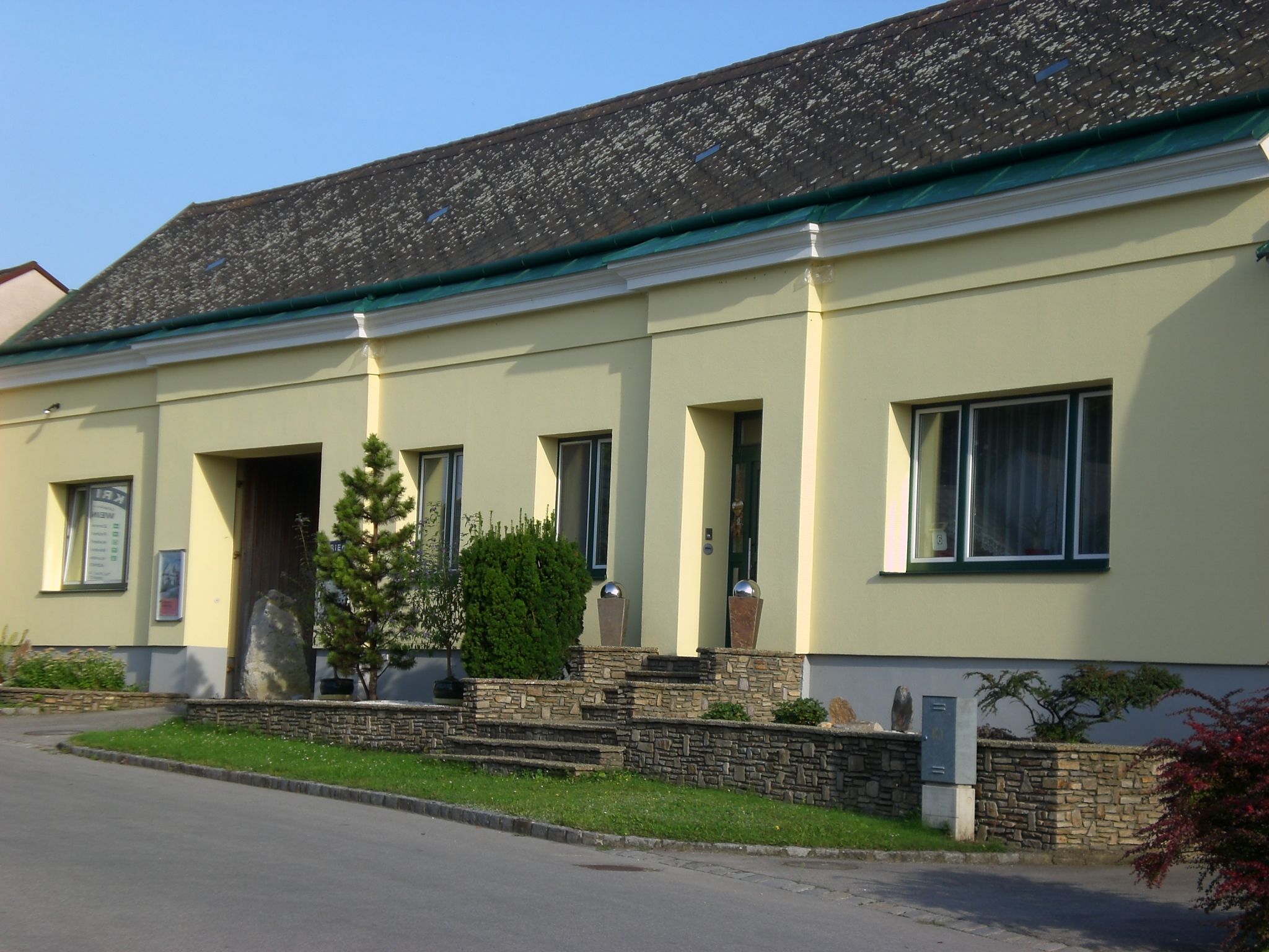 Gelbes Gebäude mit Steinmauer und Pflanzen vor dem Eingang.