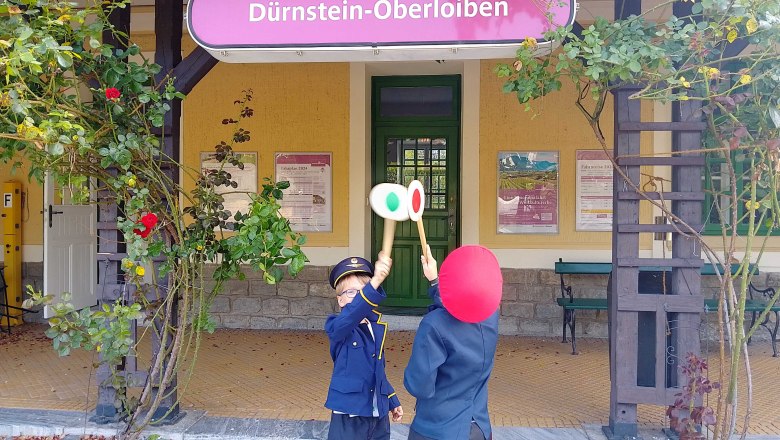 Zwei Kinder in Eisenbahnuniformen stehen vor dem Bahnhof Dürnstein-Oberloiben und halten Signaltafeln hoch.