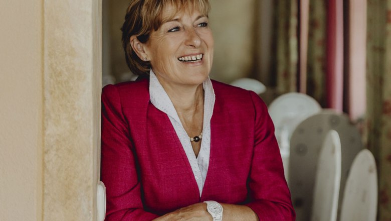 Frau in rotem Blazer lächelt und lehnt sich an eine Wand.