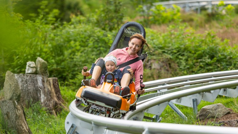 Eine Frau und ein Kind fahren auf einer Sommerrodelbahn durch eine grüne Landschaft.