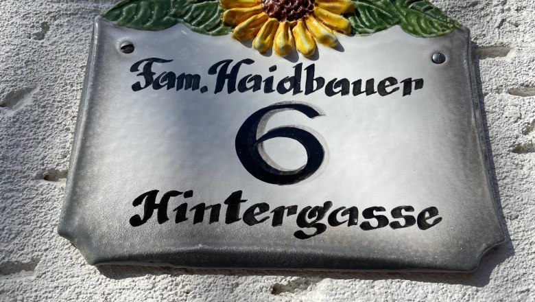 Pension Haidbauer, © Wiener Alpen Hausnummernschild mit Sonnenblume und der Aufschrift 'Fam. Haidbauer 6 Hintergasse'.
