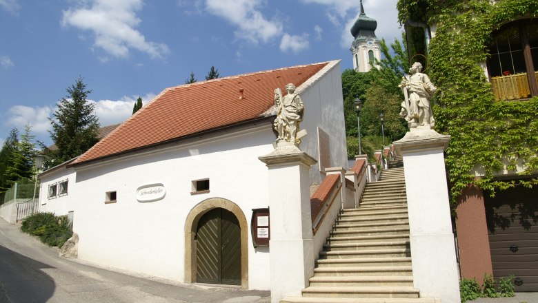 Wei&szlig;es Geb&auml;ude mit rotem Dach und Treppe mit Statuen, Kirche im Hintergrund.