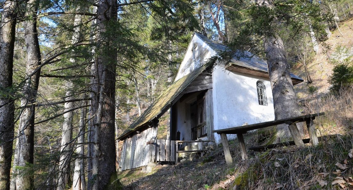 Eine kleine weiße Kapelle im Wald, umgeben von Bäumen und einem Holztisch im Vordergrund.